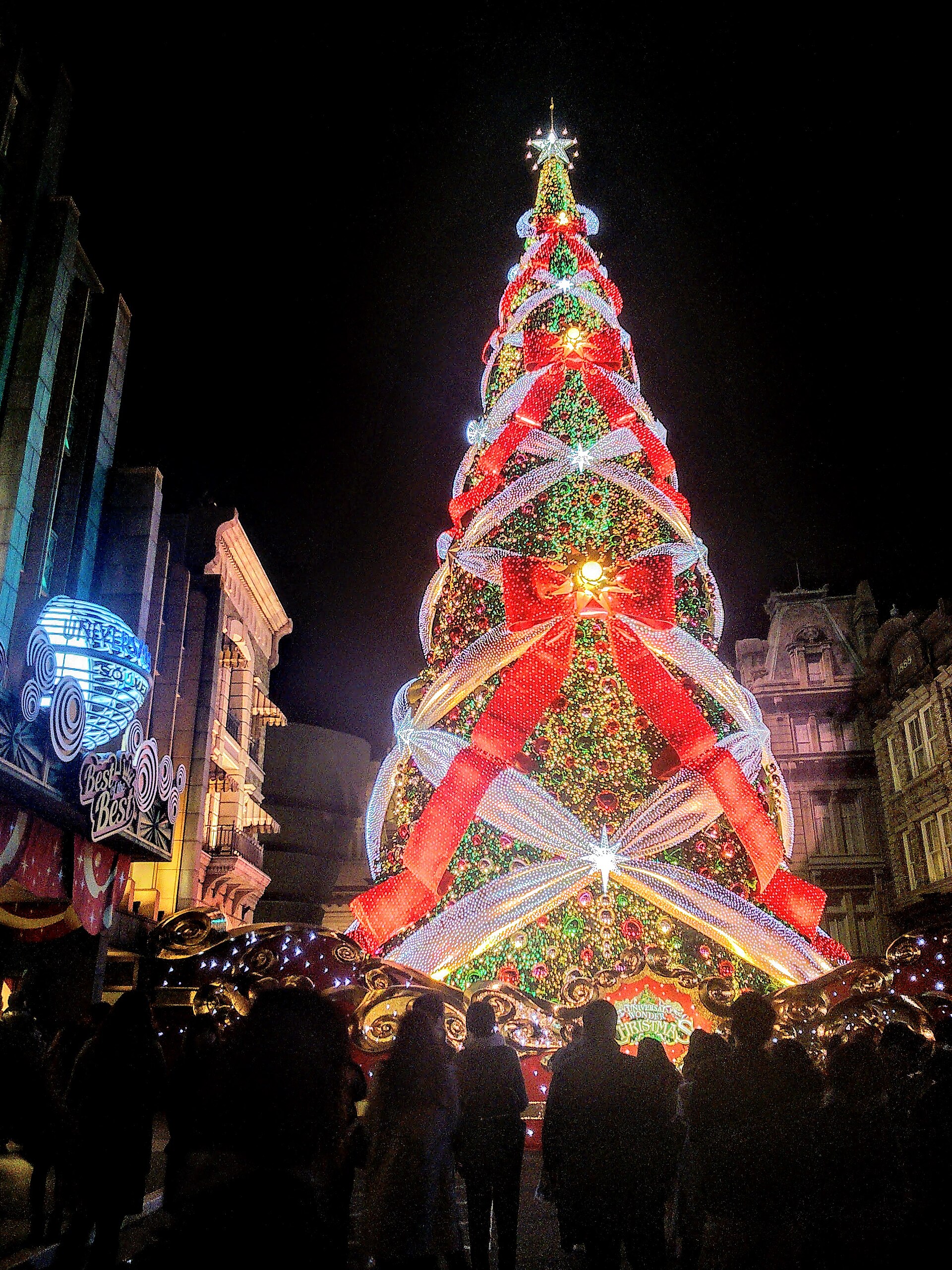 A tall Christmas tree lit in red, green and white ribbons of light in a street at Universal Studios Japan at night, with visitors gathered around its base