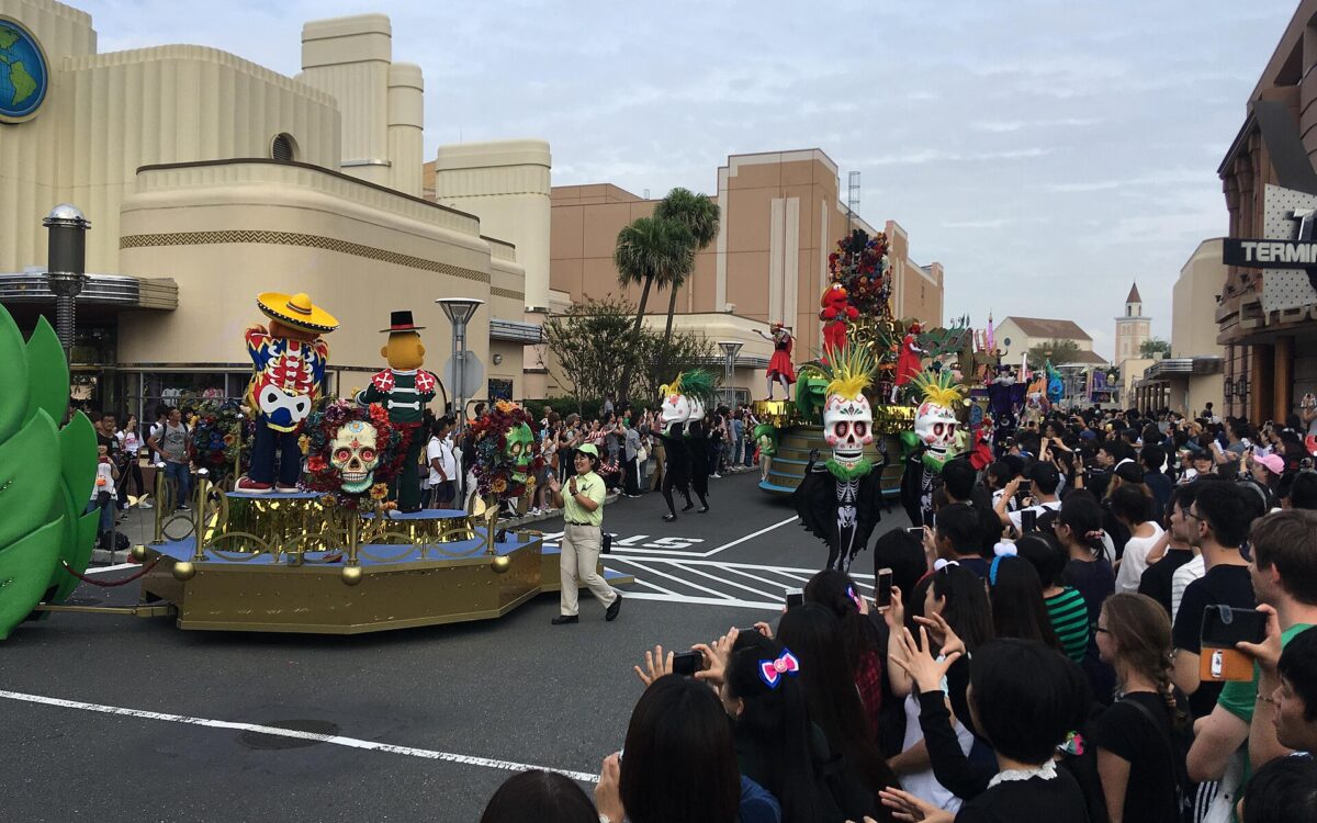A daytime parade at Universal Studios Japan, with a skull-decorated float and dancers passing a dense crowd of spectators