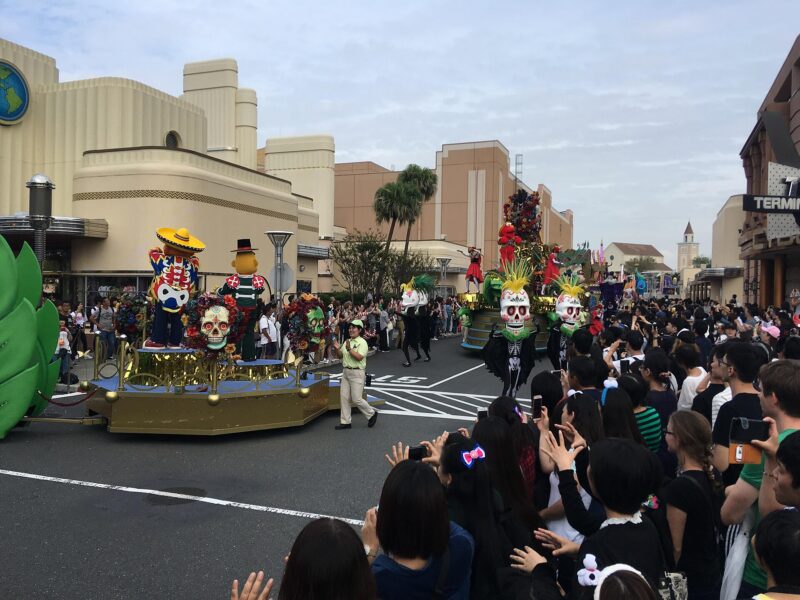 A daytime parade at Universal Studios Japan, with a skull-decorated float and dancers passing a dense crowd of spectators