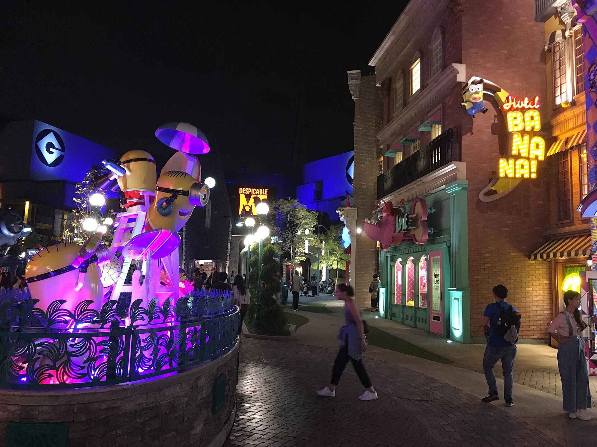 Minion Park at Universal Studios Japan after dark, with a lit-up Minion sculpture, glowing shopfronts and visitors walking along the street