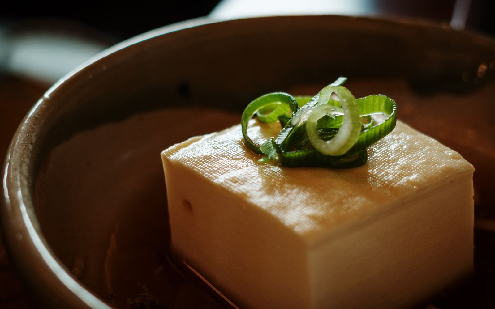A block of silken tofu topped with sliced green onion, served in a dark ceramic bowl