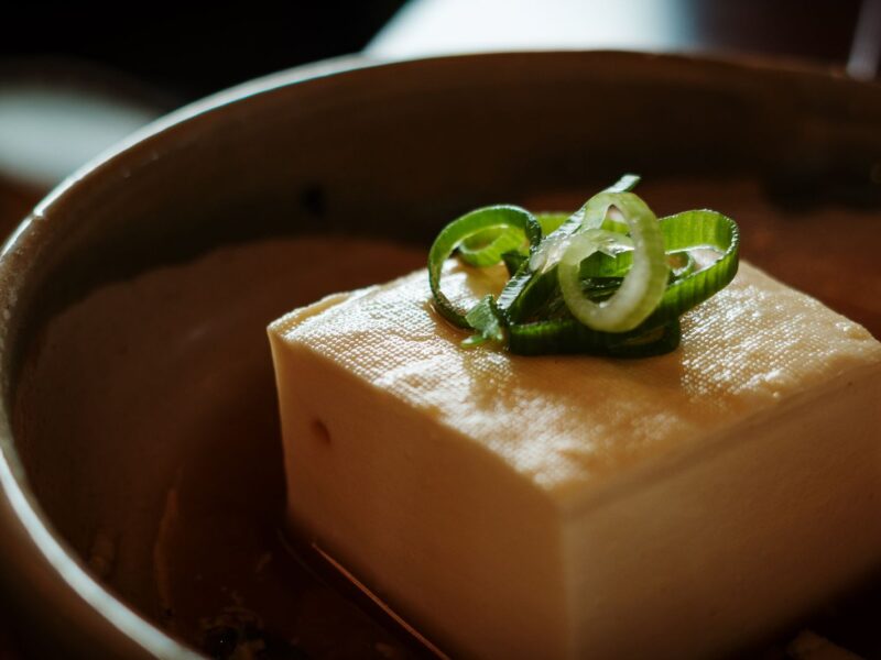 A block of silken tofu topped with sliced green onion, served in a dark ceramic bowl