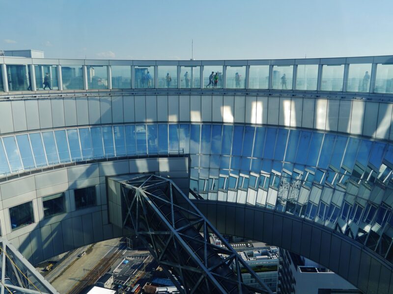 Visitors walk around the circular observation deck of Osaka's Umeda Sky Building, above an enclosed escalator and distant streets.