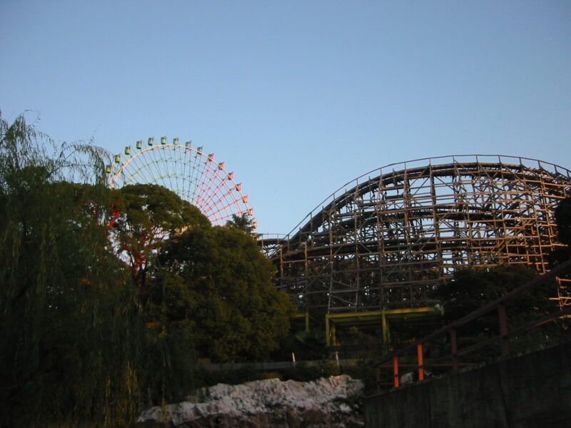 Wooden roller coaster tracks curve beside leafy trees with Hirakata Park's colorful Ferris wheel rising in the background.