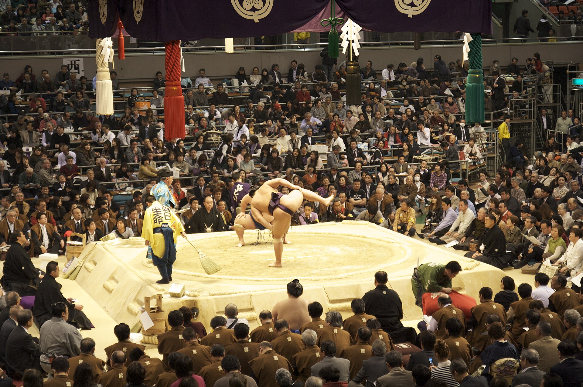 Wrestlers perform leg raises on the sumo ring at Osaka's March tournament, surrounded by seated spectators beneath coloured tassels.